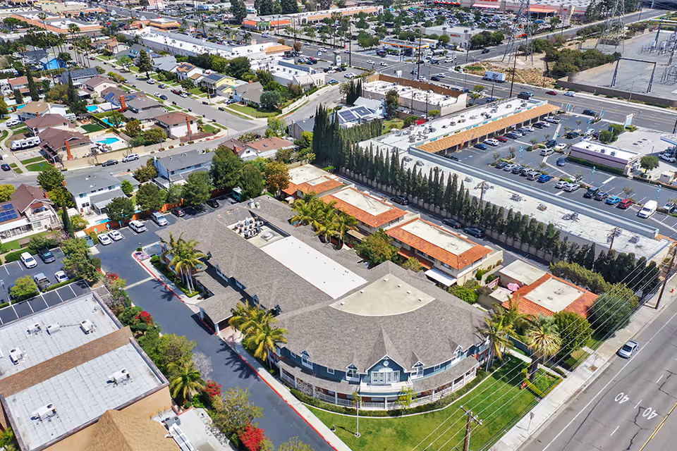 Aerial view of Kirkwood Orange facility showing a large building with a curved front, surrounded by palm trees and parking lots. Nearby are residential houses, commercial buildings, and streets with cars.