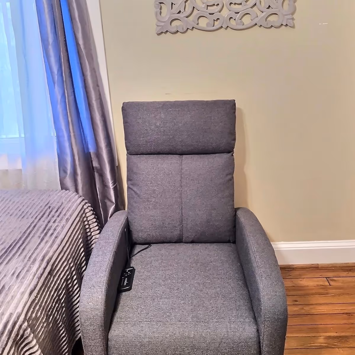 A gray upholstered recliner chair positioned next to a bed with a textured gray bedspread. The room has wooden flooring, light-colored walls, and a window with sheer white and gray curtains. A decorative white wall hanging is partially visible above the chair.