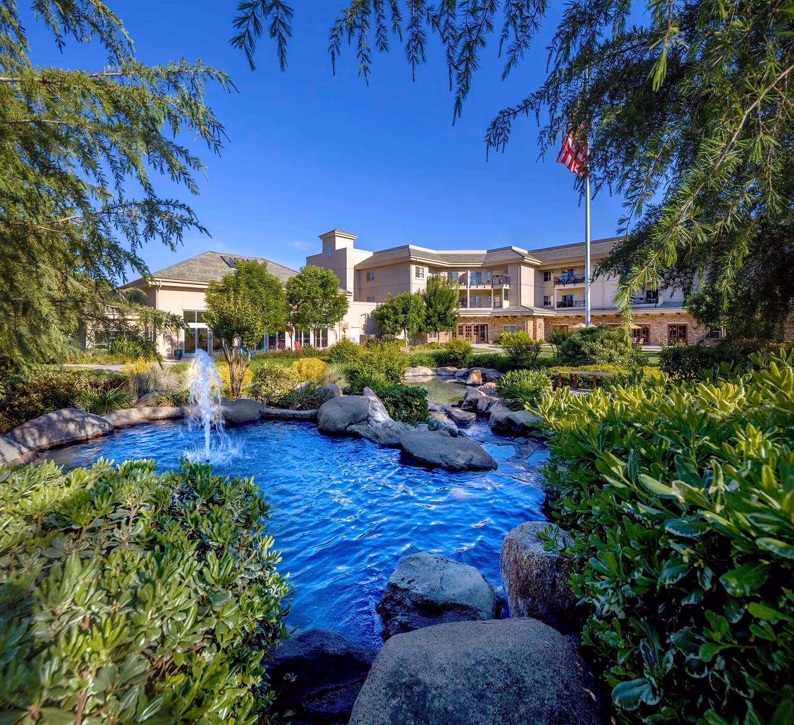 Front exterior of a senior living building with a landscaped pond and fountain surrounded by shrubs and rocks under a clear blue sky.