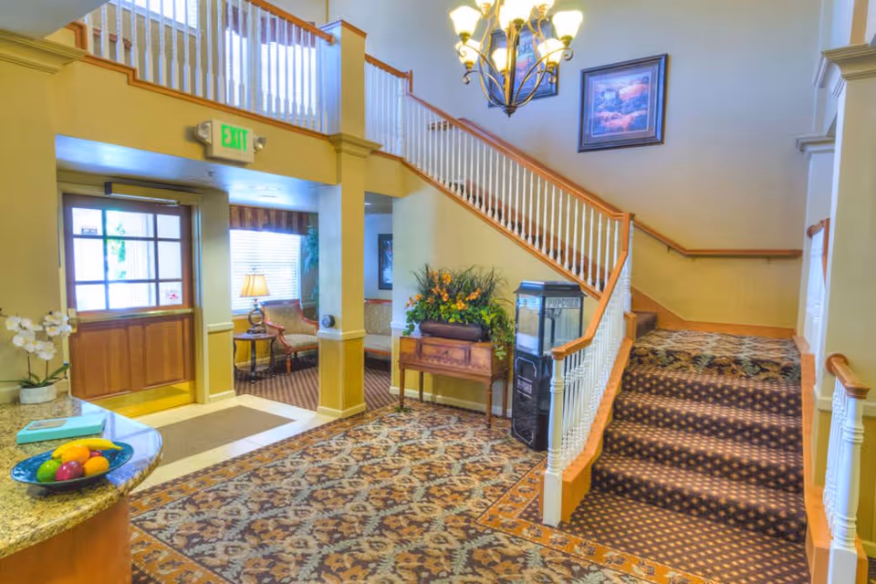 Interior view of a senior living facility lobby with a patterned carpet, a staircase with white railings and wooden handrails, a chandelier, a popcorn machine, a wooden table with a flower arrangement, and a seating area near a window with a lamp.