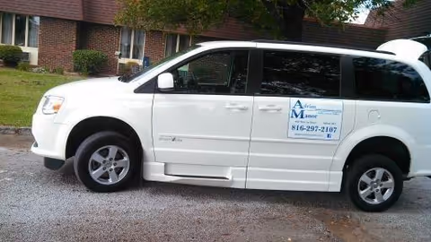 White minivan parked on a gravel driveway in front of a brick building with windows and bushes. The minivan has a sign on the side door that reads 'Adrian Manor Health & Rehab' with a phone number.