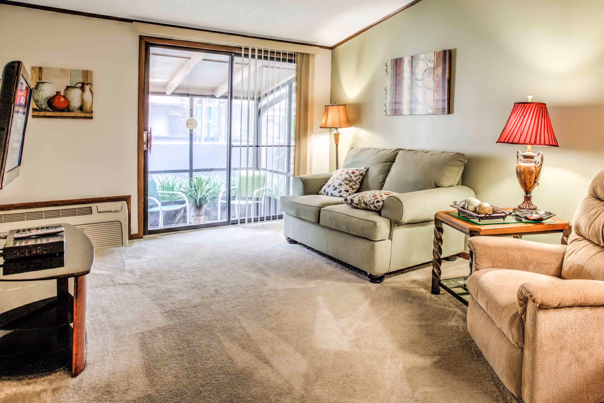 A cozy living room with a light green loveseat adorned with two patterned pillows, a beige recliner chair, and a wooden side table with a decorative lamp and ornaments. The room has beige carpet flooring and light-colored walls with two pieces of wall art. A sliding glass door with vertical blinds leads to a small outdoor patio with chairs and potted plants.