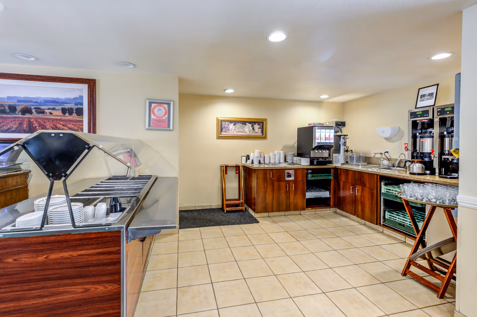 A clean and well-lit buffet area with a serving counter on the left holding plates and cups under a transparent sneeze guard. On the right side, there is a countertop with coffee machines, stacks of cups, glasses, and a sink. The walls are decorated with framed pictures, and the floor is tiled.