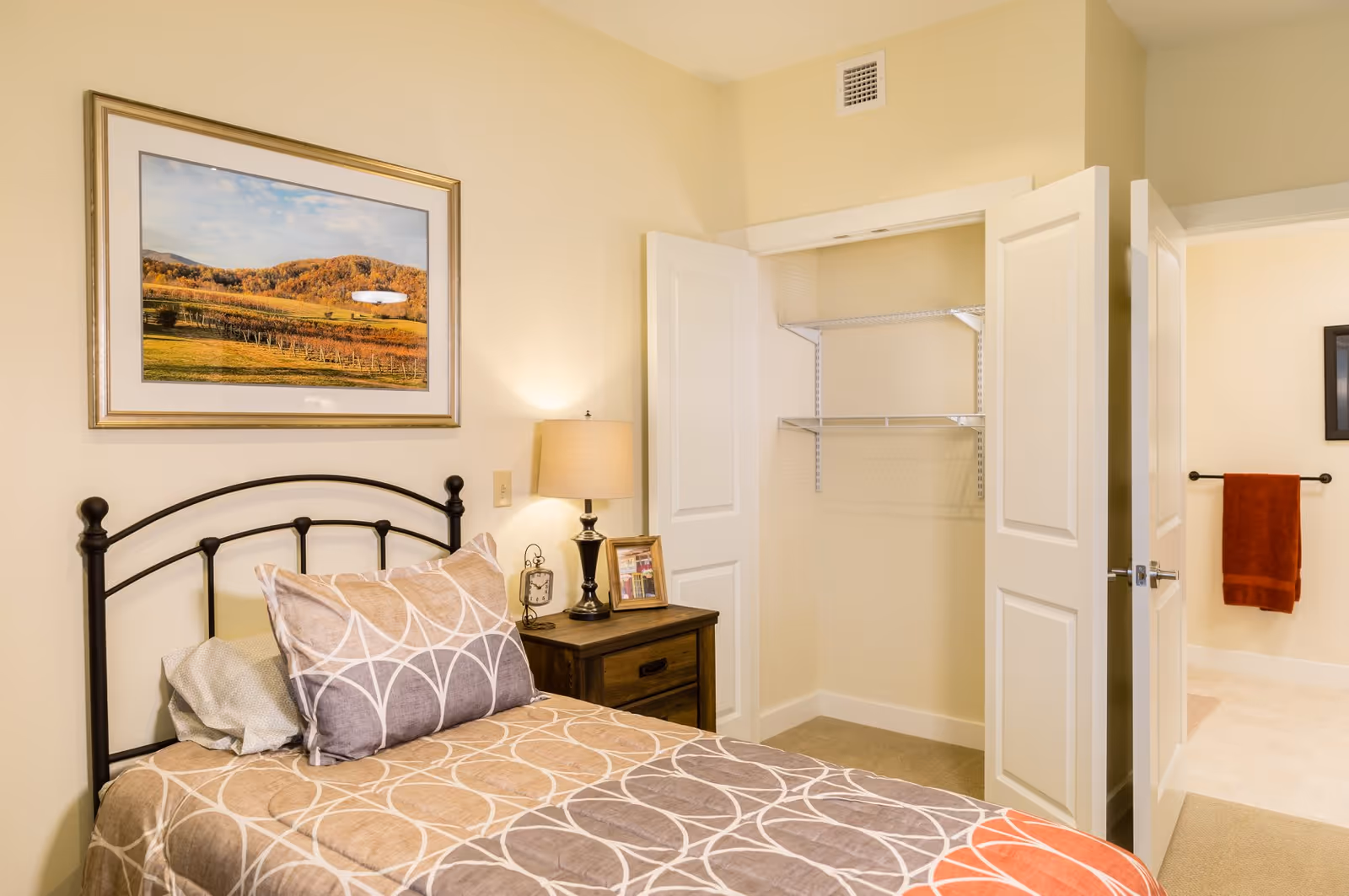 A cozy bedroom in a senior living facility featuring a single bed with patterned bedding, a wooden nightstand with a lamp, clock, and framed photo, an open closet with white double doors, and a framed landscape picture hanging on the wall above the bed.