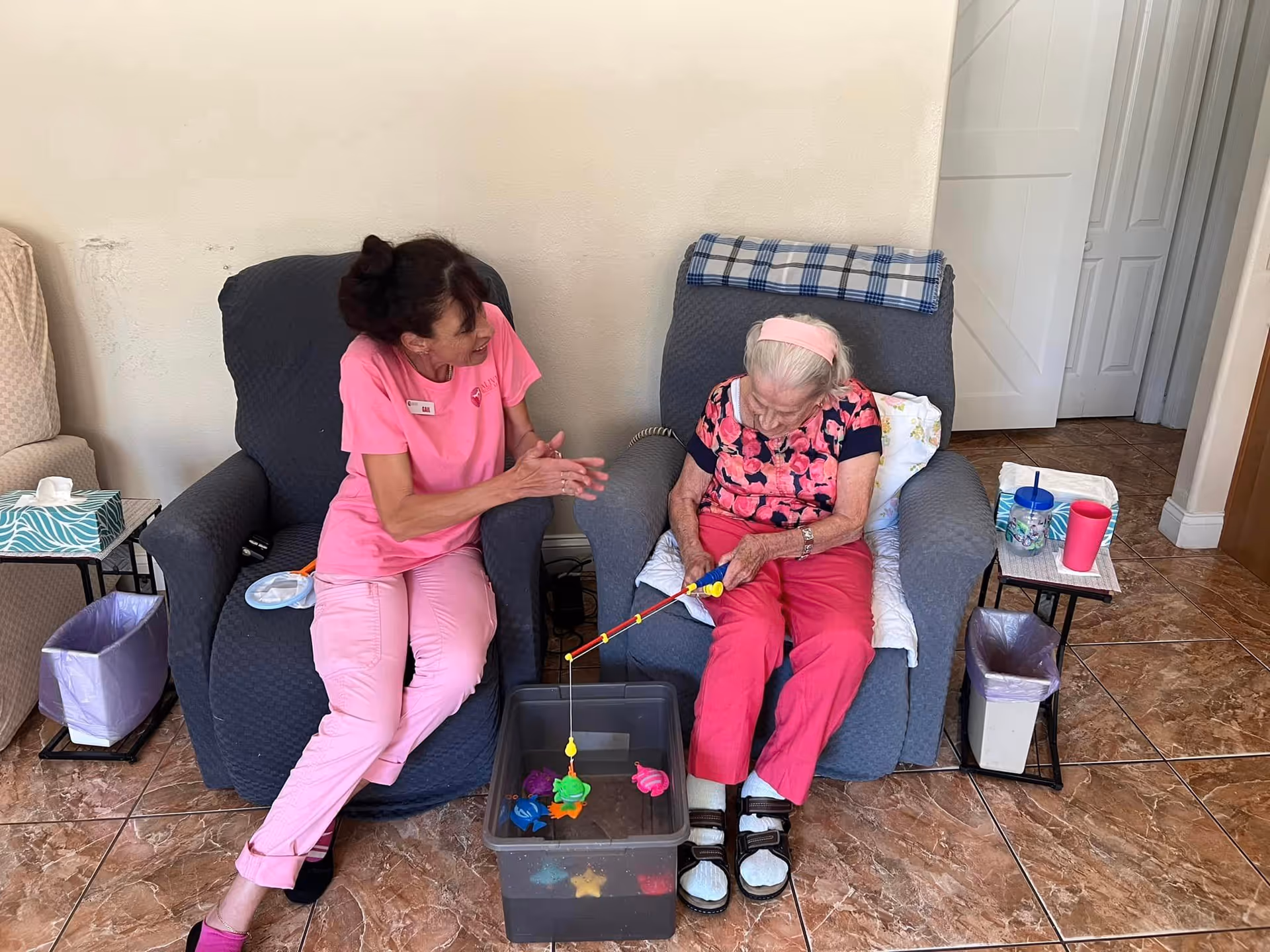 An elderly woman sitting in a blue armchair playing with a fishing toy in a plastic container filled with water and colorful floating toys, accompanied by a caregiver in pink scrubs sitting next to her and smiling. The room has tiled floors, two small side tables with cups and tissues, and a beige wall in the background.