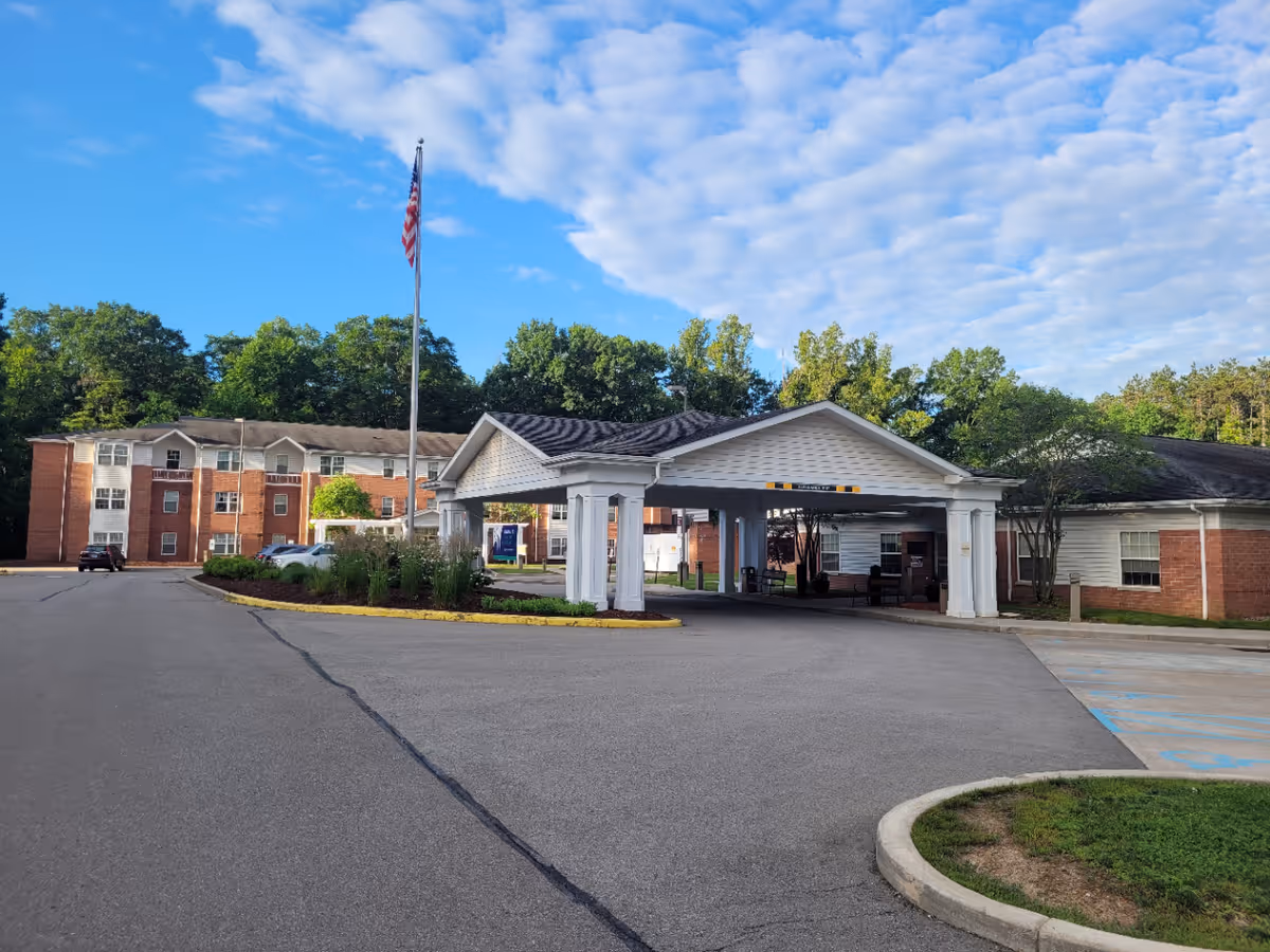 Exterior view of Jameson Nursing and Rehabilitation facility showing a covered entrance with white pillars, a flagpole with an American flag, a parking area, and a multi-story brick building surrounded by trees under a partly cloudy blue sky.