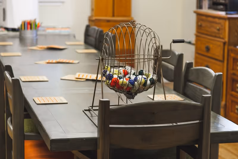 A long wooden table with multiple chairs around it, set up for a bingo game with bingo cards and a metal cage containing numbered bingo balls in the center. The room has wooden furniture and a cozy atmosphere.