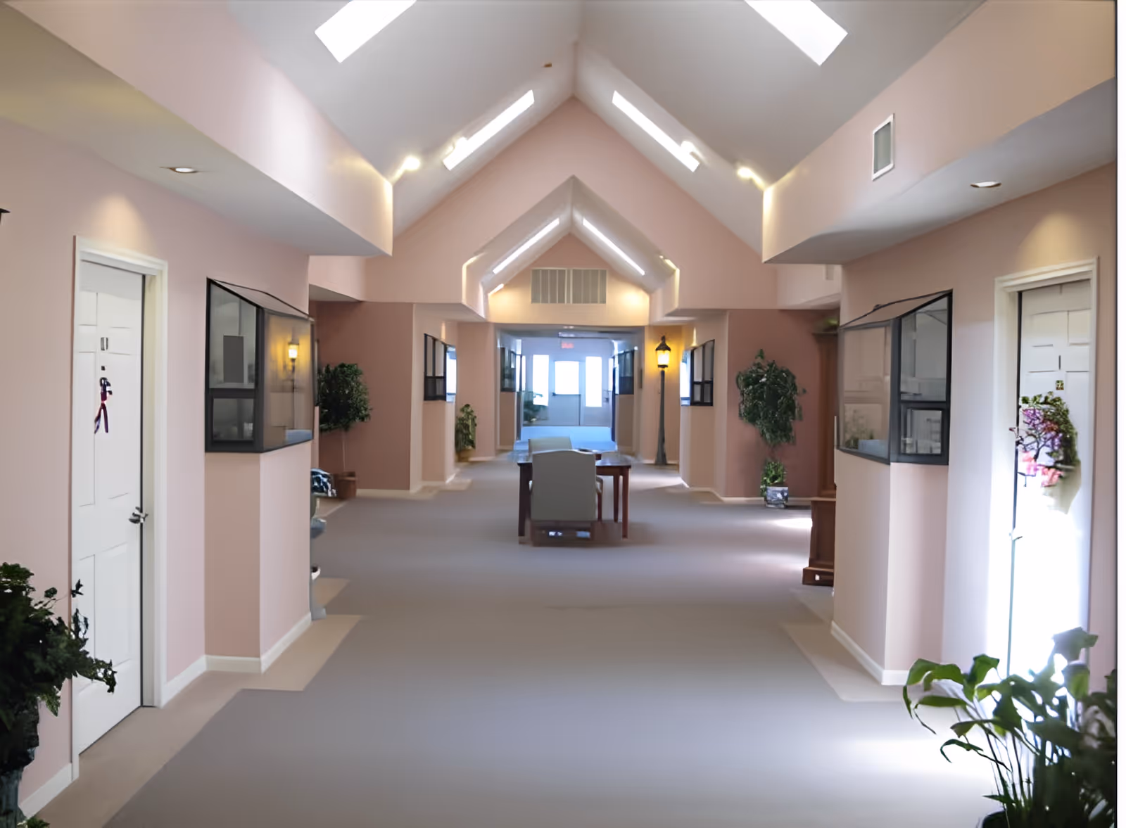 A bright, spacious hallway in an assisted living facility with soft pink walls and a high vaulted ceiling featuring skylights. The hallway has white doors on both sides, some decorated with small wreaths or ornaments. There are plants placed along the walls and a table with chairs in the middle distance. The far end of the hallway is illuminated by natural light from windows.