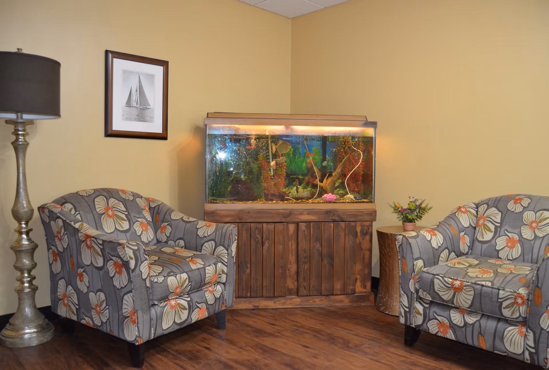 A cozy sitting area with two floral patterned armchairs facing each other, a tall metallic floor lamp on the left, a wooden cabinet with a large fish tank on top in the center, and a small round wooden side table with a small flower pot on the right. A framed black and white picture of a sailboat hangs on the beige wall behind the left armchair.