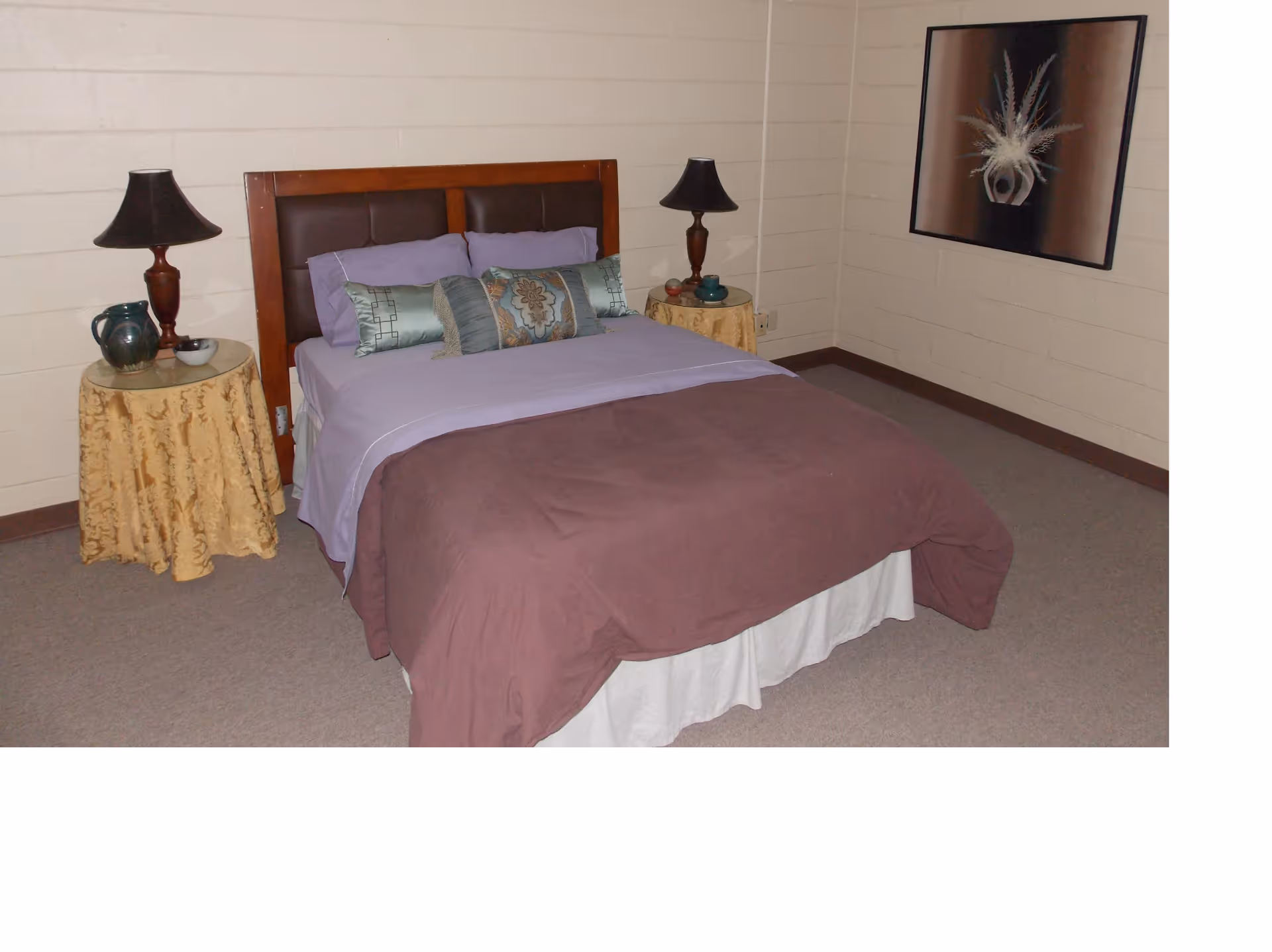 A bedroom with a double bed featuring a wooden headboard and purple bedding, flanked by two round side tables covered with yellow patterned cloths. Each table has a dark lamp and decorative items. A framed artwork hangs on the wall to the right.