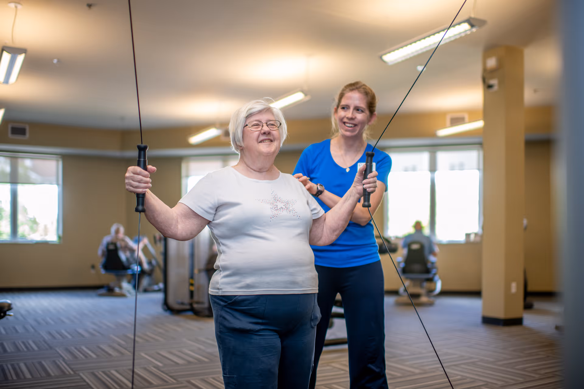 An elderly woman is exercising with resistance cables in a fitness room, assisted by a younger female instructor who is smiling and providing support. The room has large windows, beige walls, and exercise equipment in the background.