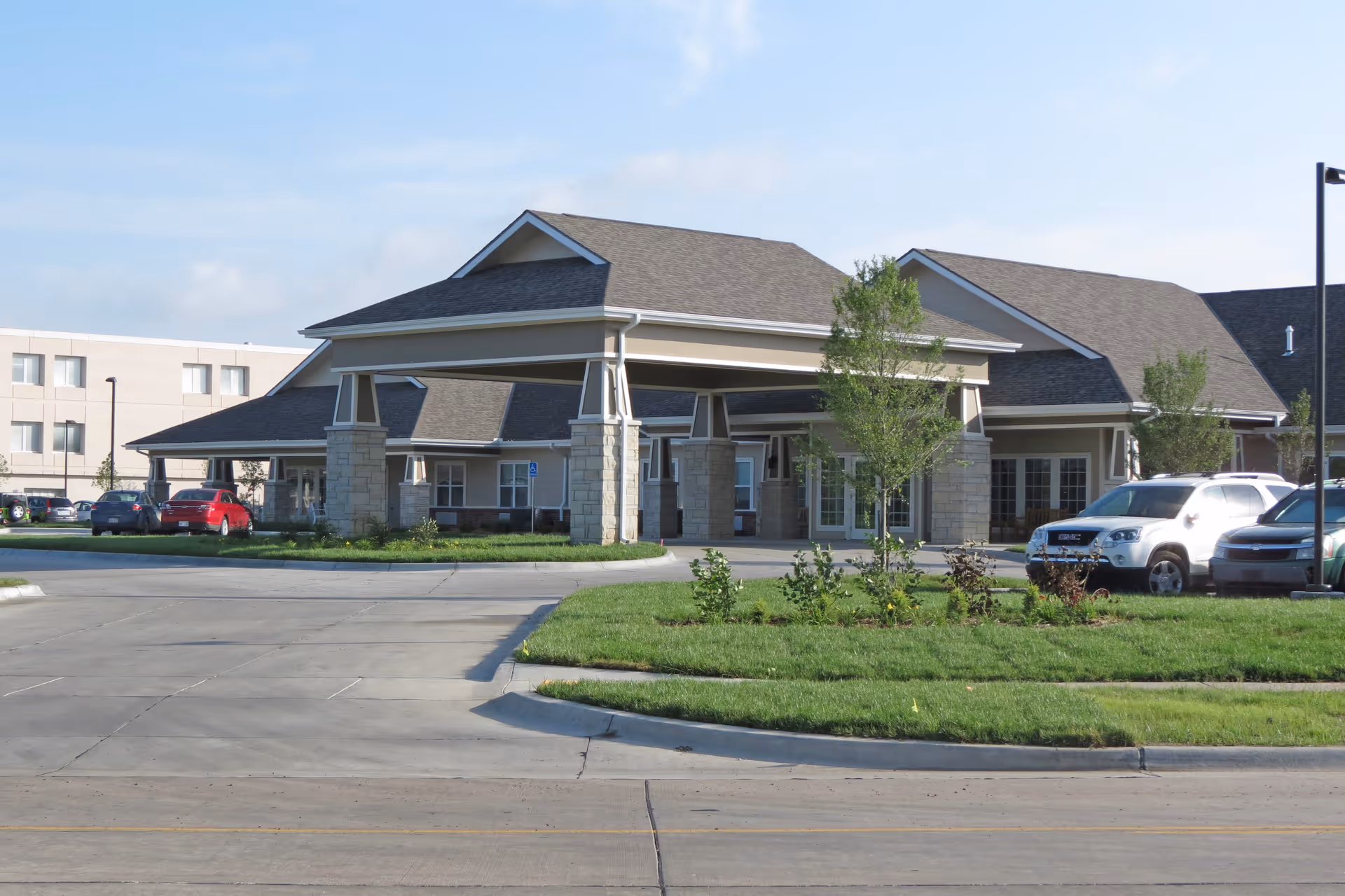 Exterior view of a single-story building with a covered entrance supported by stone pillars, surrounded by a parking lot with several cars and landscaped greenery under a clear sky.