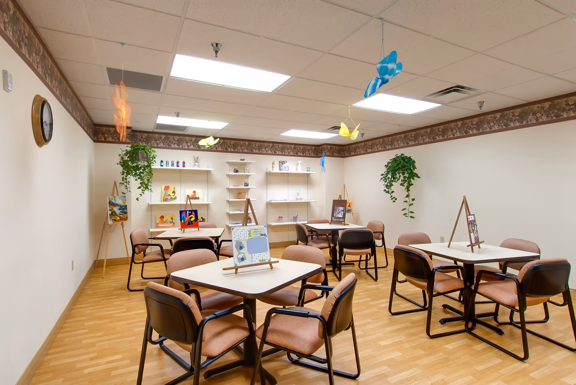 A well-lit activity room with four square tables, each surrounded by four chairs. The tables display small easels with artwork. The room has light-colored walls with a floral border near the ceiling, hanging plants, and colorful butterfly decorations suspended from the ceiling. Shelves on the far wall hold various decorative items and art supplies.
