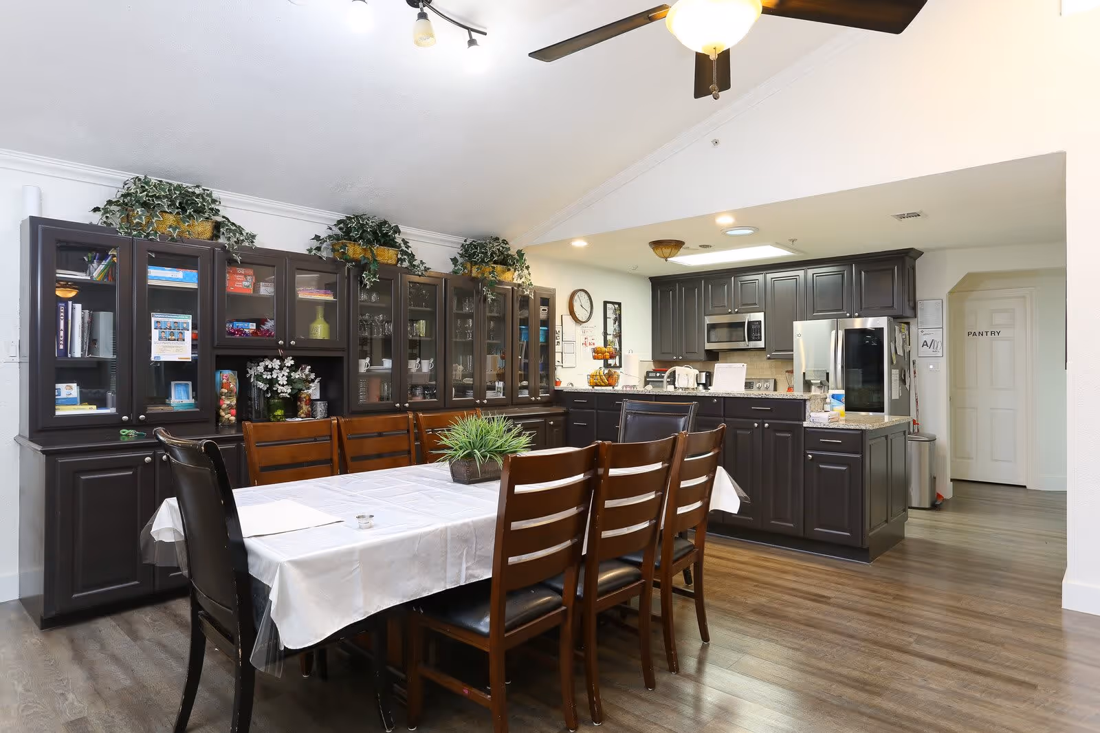 A dining area with a long table covered with a white tablecloth surrounded by wooden chairs with black cushions. Behind the table, there is a large dark wood cabinet with glass doors displaying books, decorative items, and glassware. To the right, there is a kitchen area with dark cabinetry, a stainless steel refrigerator, microwave, and various kitchen appliances. The floor is wooden, and the ceiling has a fan with lights. A door labeled 'PANTRY' is visible in the background.