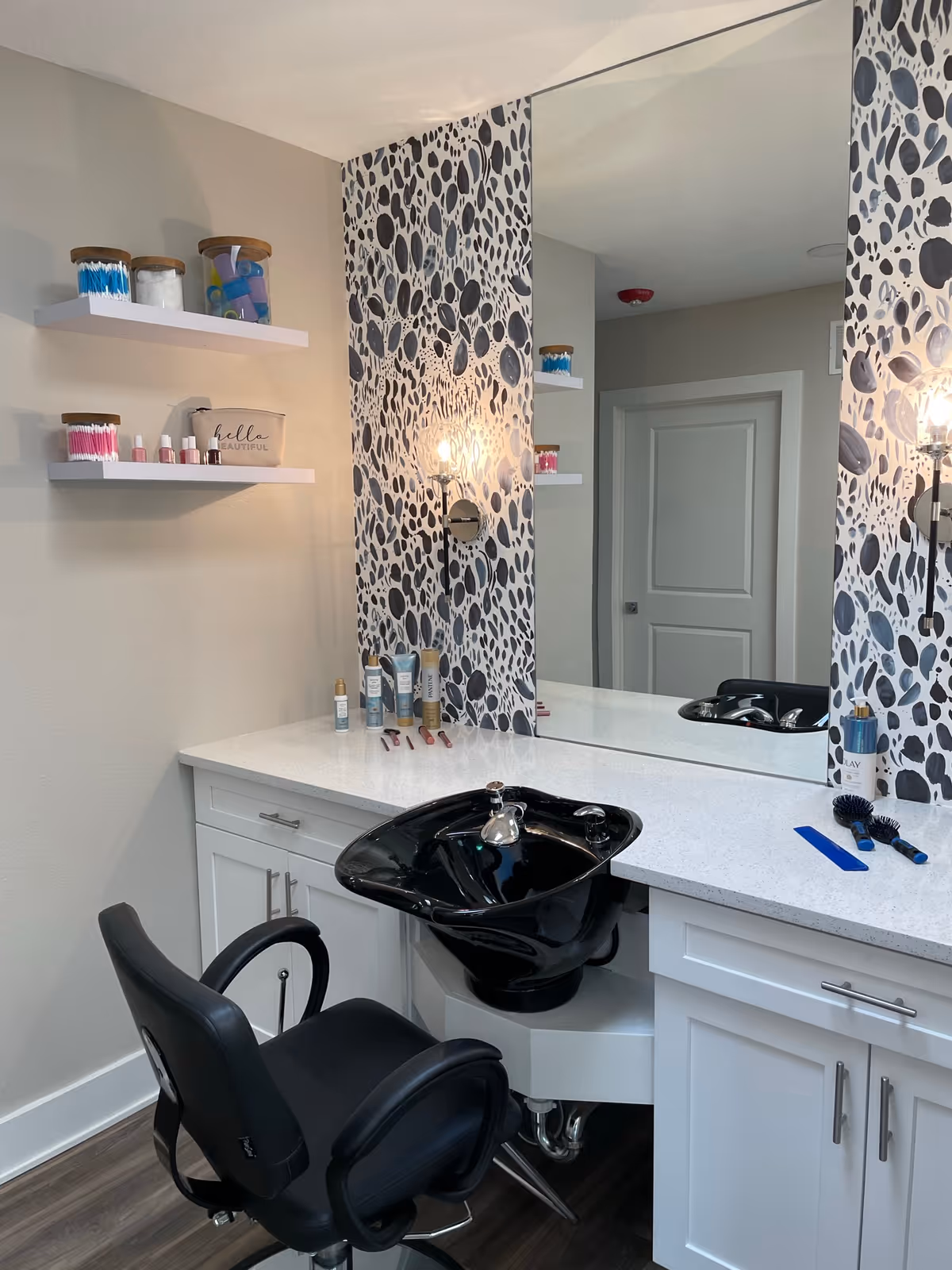 A salon station with a black salon chair and a black shampoo sink set into a white countertop. The wall behind features a large mirror and a decorative panel with a black and white abstract pattern. Two white floating shelves hold various beauty supplies, including cotton swabs and nail polish. A wall-mounted light fixture with an exposed bulb is also visible.