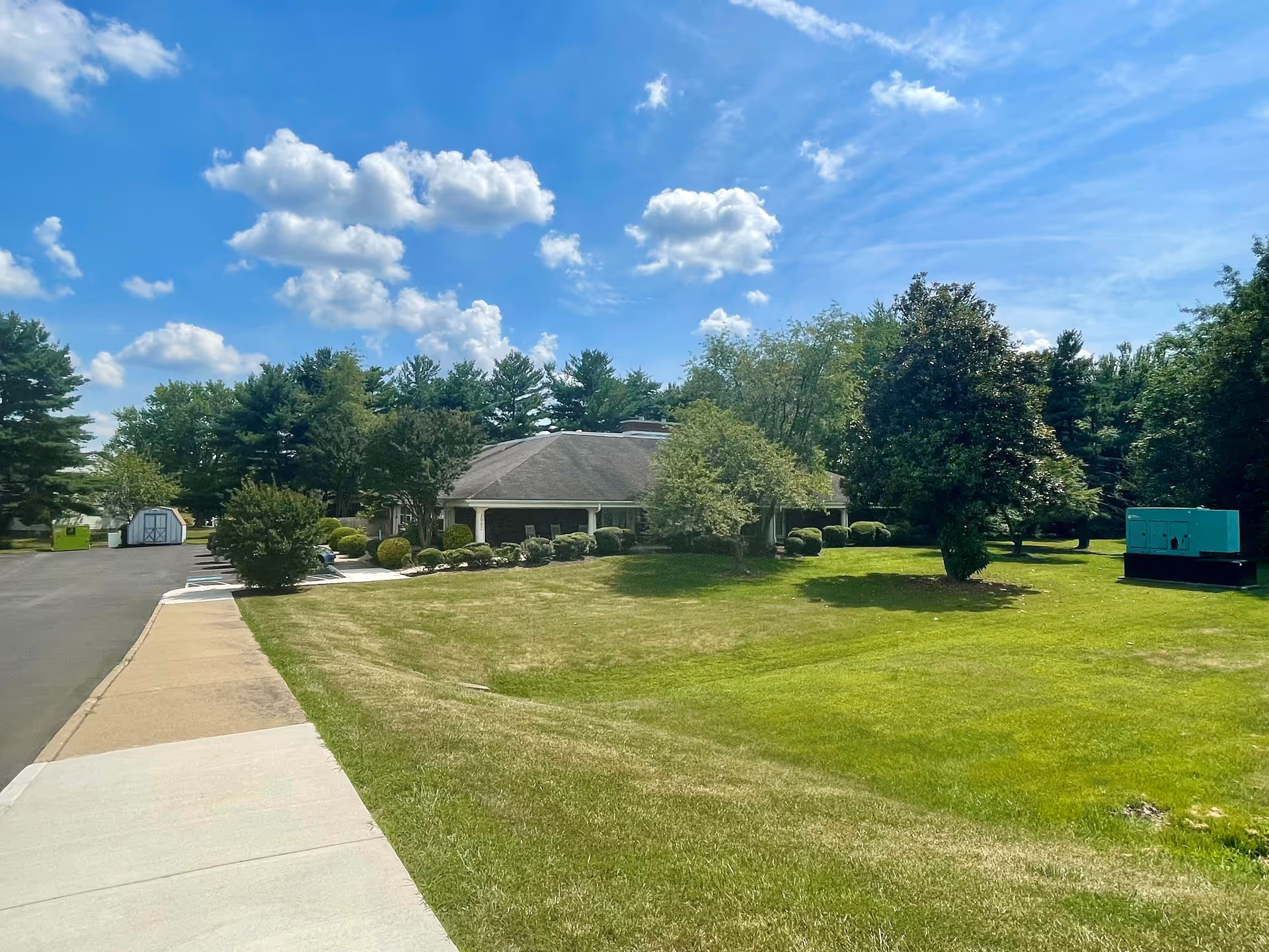 Single-story assisted living building with a wraparound porch set amid a large grassy lawn and trees under a blue sky.