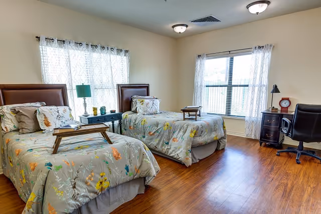 A bright bedroom with two twin beds featuring floral bedspreads and wooden headboards. Each bed has a wooden tray on top. Between the beds is a small table with a green lamp and decorative items. There is a large window with sheer white curtains letting in natural light. On the right side of the room, there is a black office chair and a small dark wooden nightstand with a red clock and a lamp. The floor is wooden, and the walls are painted a light beige color.