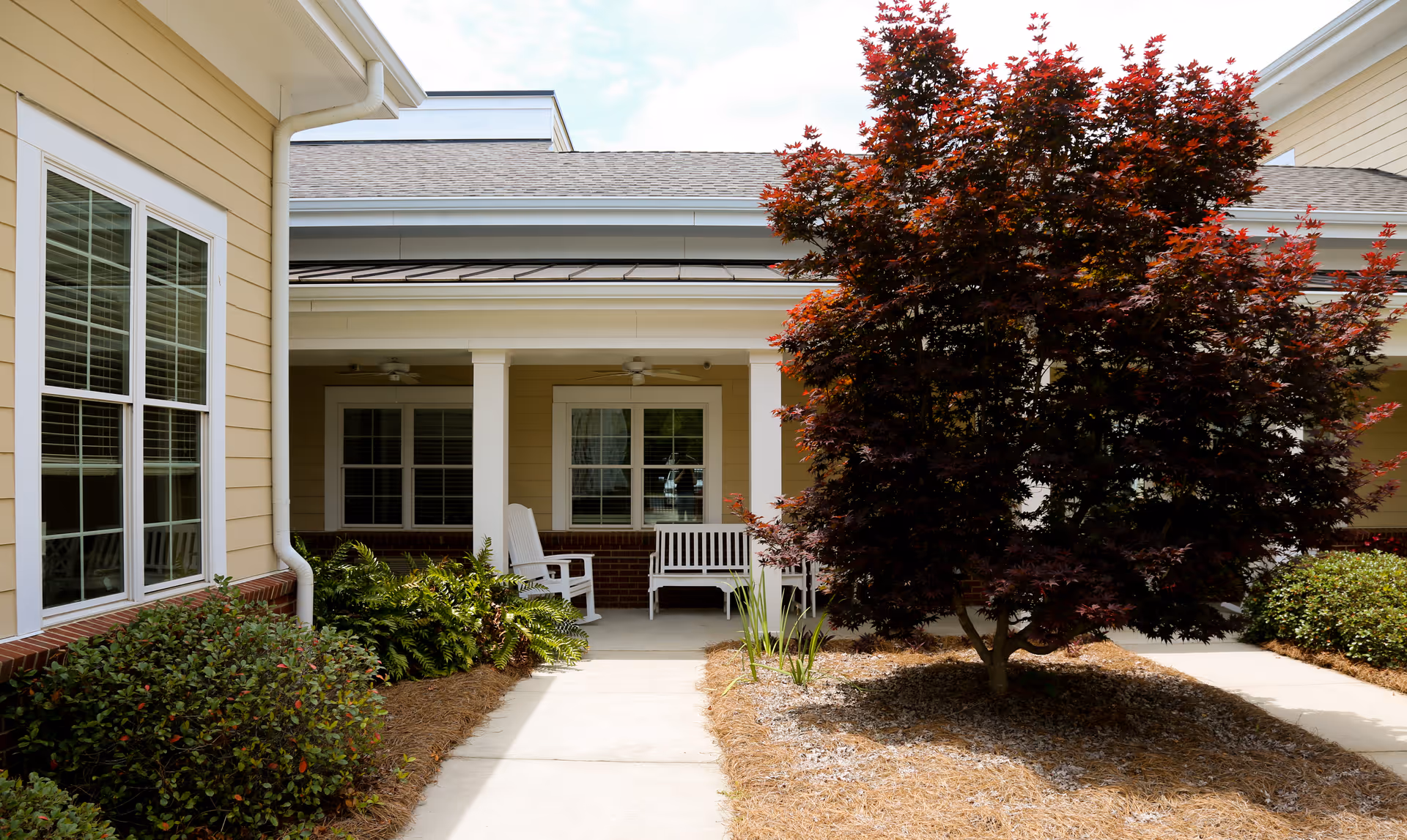 Outdoor patio area at Zebulon Park Health & Rehabilitation featuring a covered seating area with white chairs and a bench, surrounded by bushes and a tree with dark red leaves.