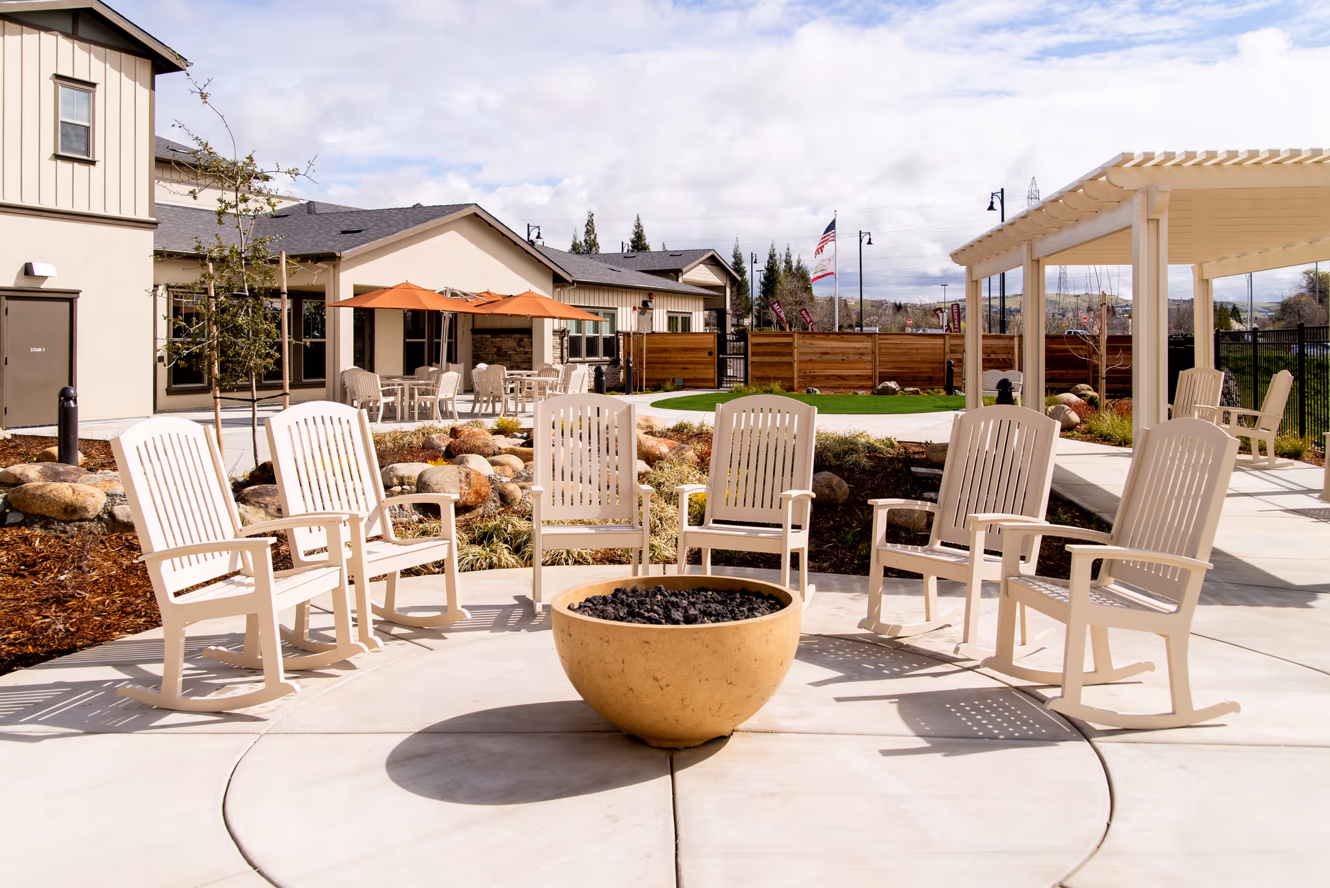 Outdoor seating area at CountryHouse Residence featuring six beige rocking chairs arranged in a circle around a large fire pit. The area is paved with concrete and surrounded by landscaping with rocks and plants. In the background, there are buildings with beige walls, tables with orange umbrellas, a pergola, and a wooden fence. The sky is partly cloudy.