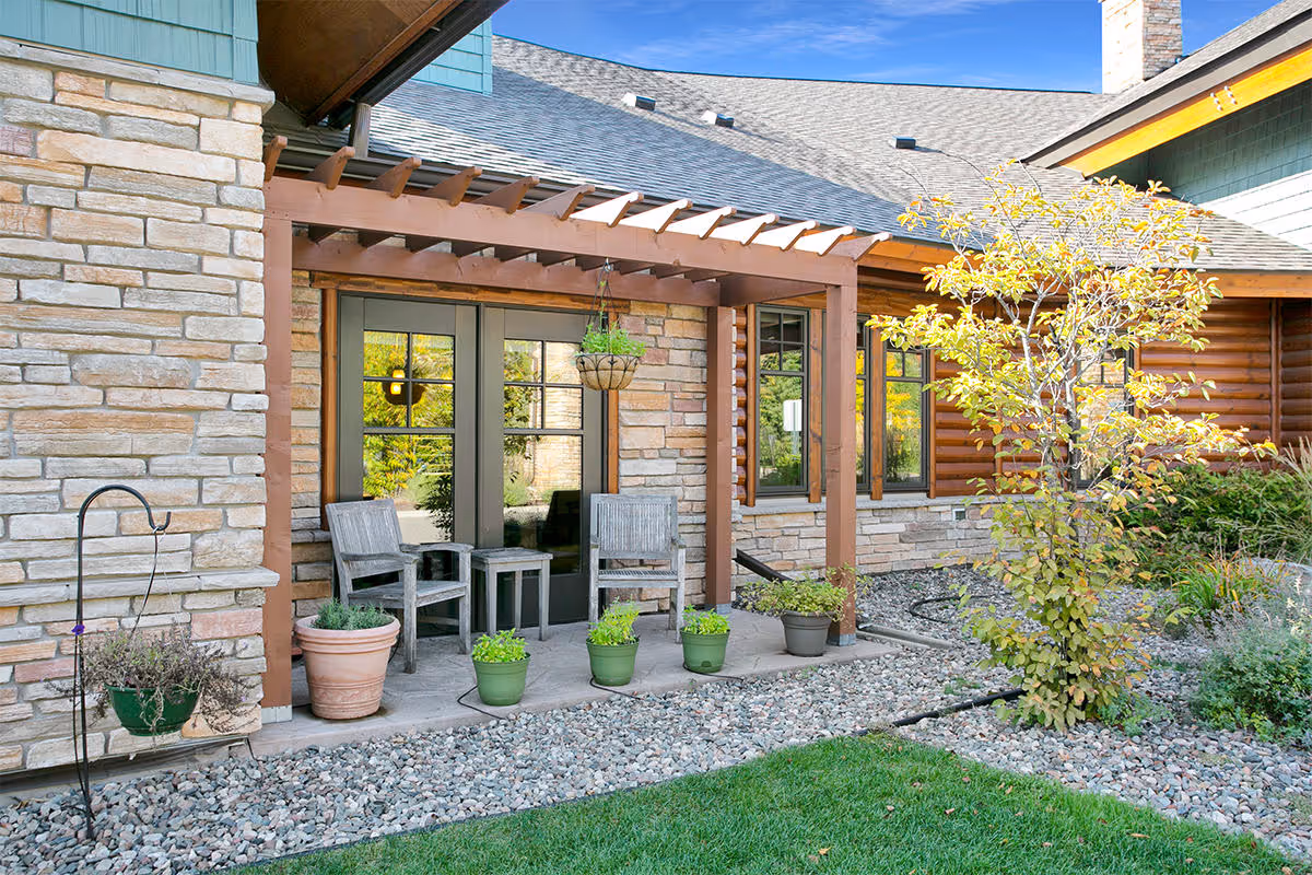 Outdoor patio area with two wooden chairs and a small table under a wooden pergola attached to a stone and wood exterior building. Several potted plants and a small tree are visible, along with a gravel bed and green grass in the foreground.