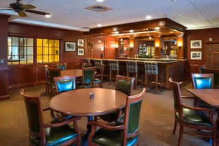 A cozy dining area with round wooden tables and green cushioned chairs. In the background, there is a bar with high stools and warm lighting, wood-paneled walls, and framed pictures hanging on the walls.
