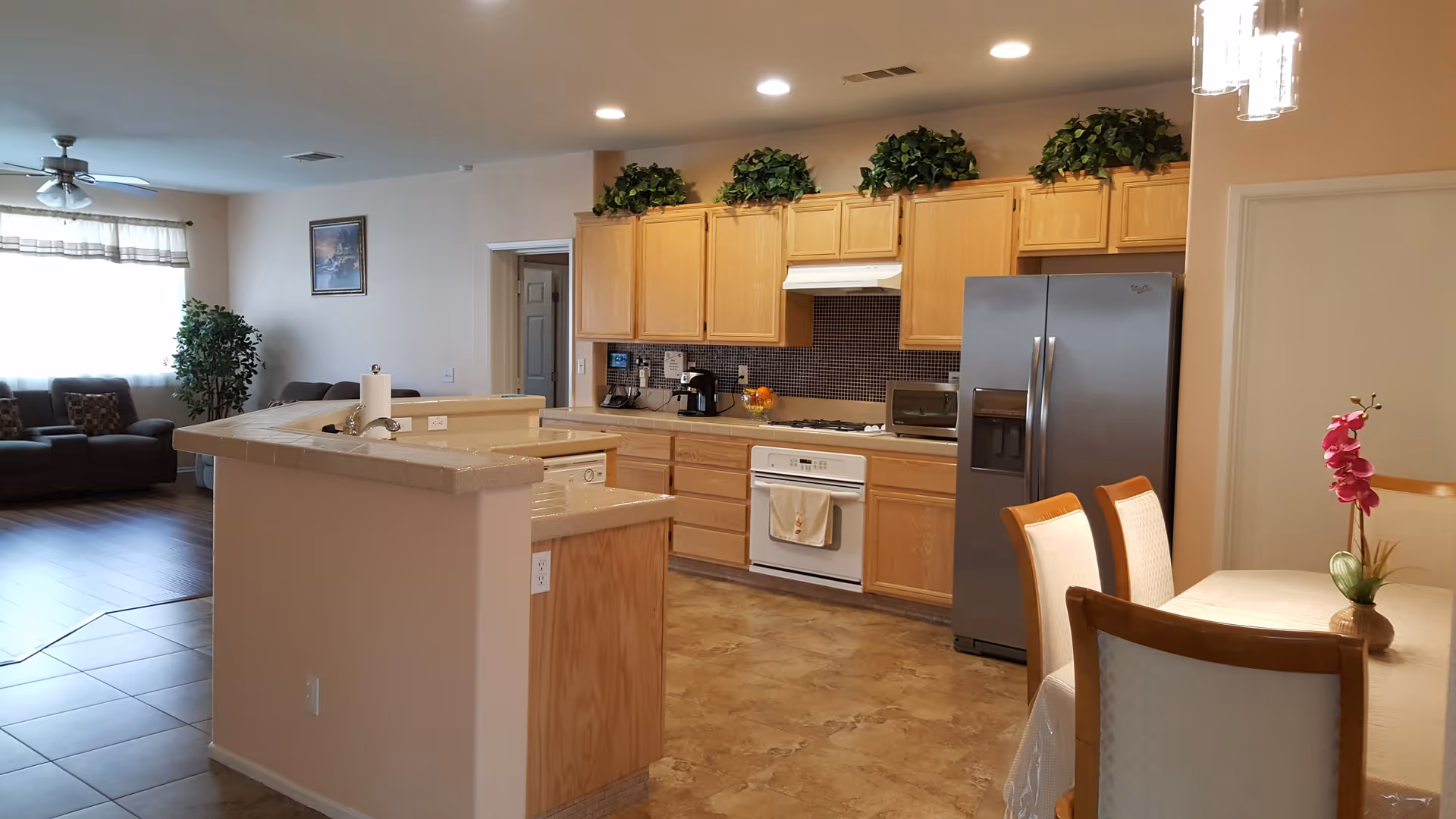 A spacious kitchen and dining area in an elderly care facility. The kitchen features wooden cabinets with green plants on top, a stainless steel refrigerator, a white stove with a range hood, a microwave, and a coffee maker. There is a kitchen island with a sink and paper towel holder. Adjacent to the kitchen is a dining table with four chairs and a pink orchid centerpiece. In the background, a living room area with sofas, a large window with curtains, a ceiling fan, and a potted plant is visible.