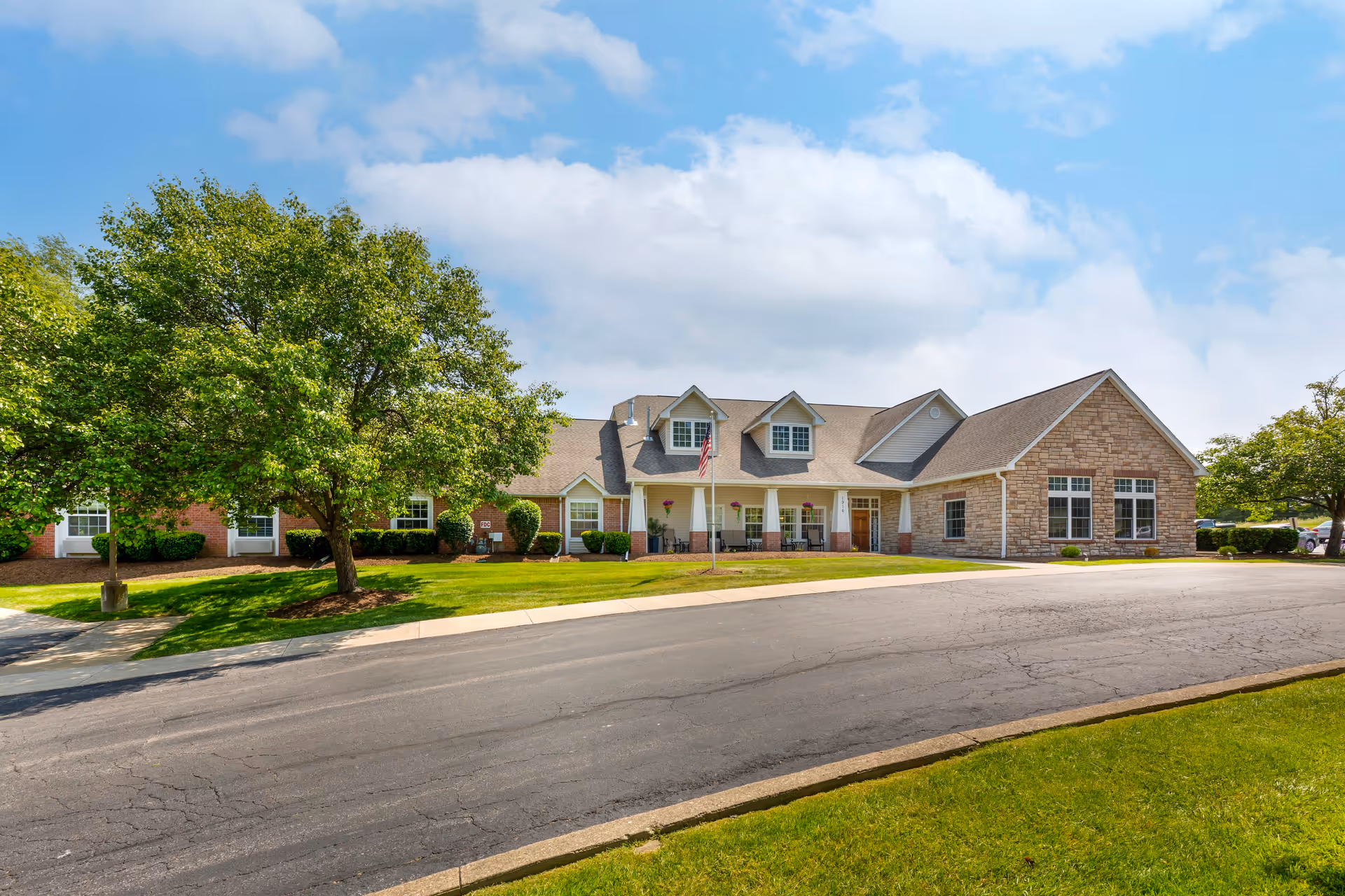 Exterior view of a single-story senior living facility building with a sloped roof, dormer windows, and a covered porch area. The building is surrounded by green lawns, trees, and a paved driveway under a partly cloudy blue sky.