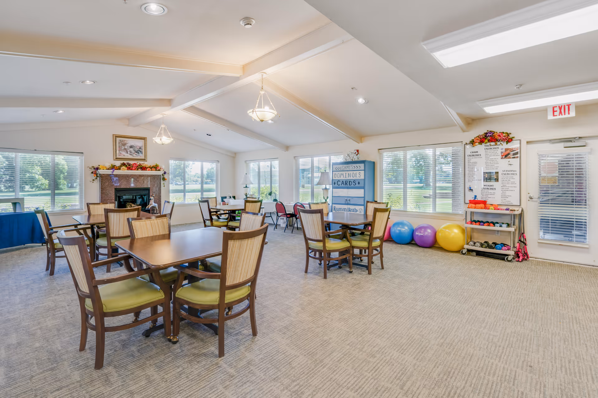A bright and spacious common room with multiple tables and chairs arranged for group activities. Large windows let in natural light and offer views of greenery outside. There is a fireplace decorated with flowers, a board listing games like dominoes and cards, colorful exercise balls, and a rack with small weights. The room has a carpeted floor and ceiling lights, with an exit door on the right side.