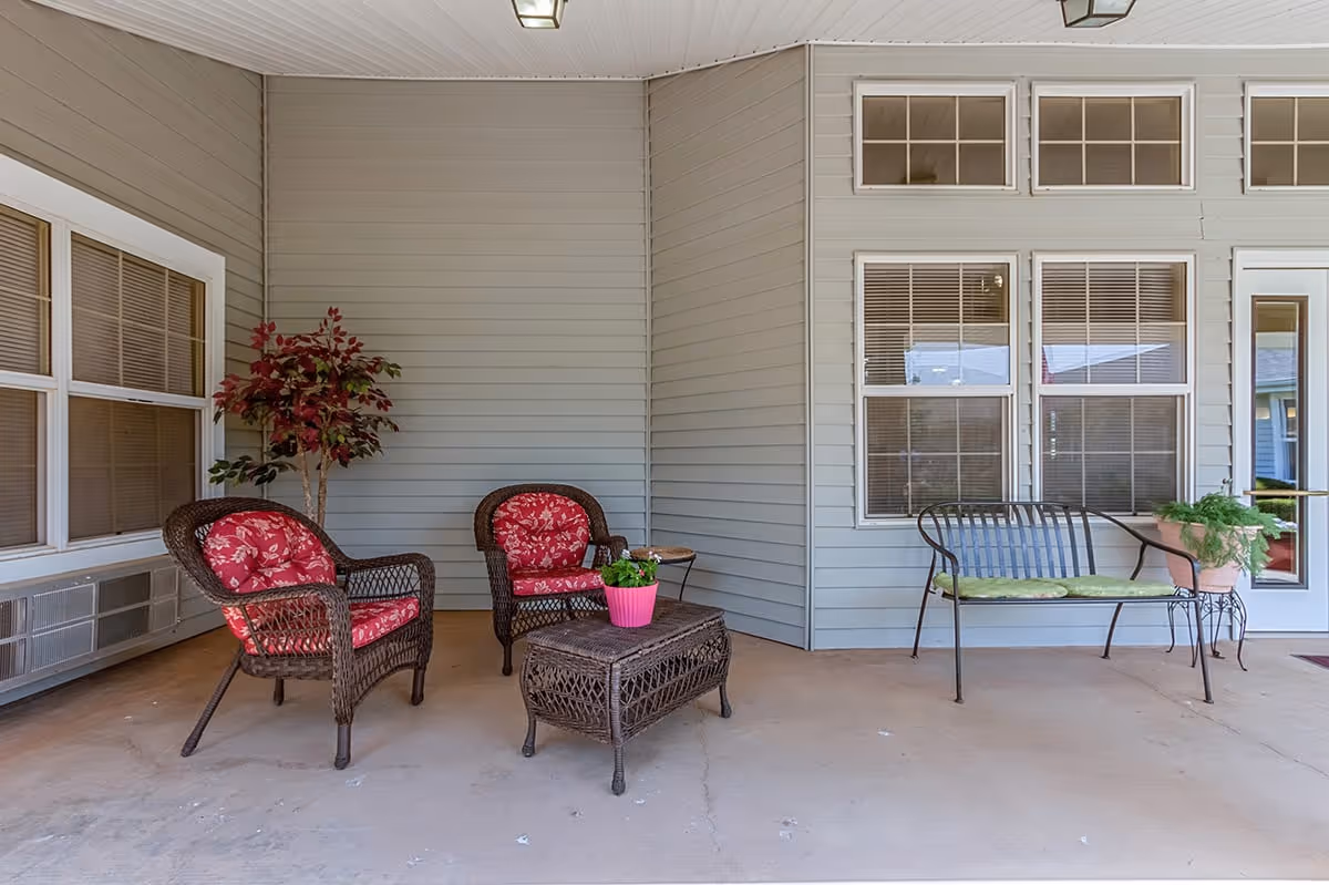 Covered outdoor patio area with two wicker chairs featuring red floral cushions, a wicker coffee table with a pink flower pot, a metal bench with a green cushion, and potted plants near the windows and door of a building.