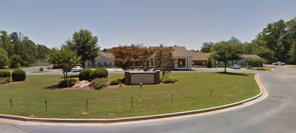Front exterior view of Stone Brooke Suites, a single-story building with a large green lawn, trees, bushes, and a sign in the center. Several cars are parked near the building under a clear blue sky.