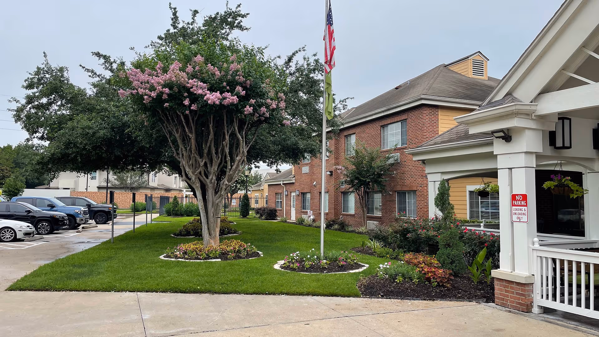 Exterior view of Atria Westchase facility showing a well-maintained lawn with flower beds and a tree with pink blossoms. An American flag is raised on a flagpole near the building entrance. Several parked cars are visible on the left side, and the building features brick and yellow siding with multiple windows. A covered porch area with hanging plants and a 'No Parking Loading & Unloading Only' sign is also visible.