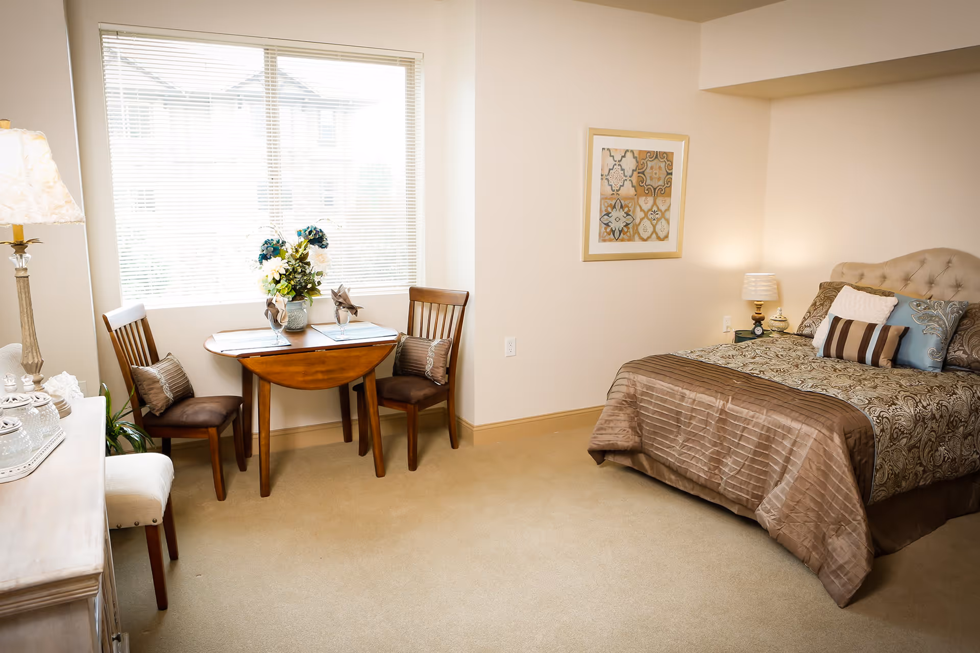 A cozy bedroom with a neatly made bed featuring patterned bedding and multiple pillows. To the left of the bed is a small wooden table with two chairs, decorated with a vase of flowers and placemats. A large window with blinds allows natural light to fill the room. A framed piece of artwork hangs on the wall above the bed, and a lamp sits on a nightstand beside the bed. The room has beige carpeting and light-colored walls.