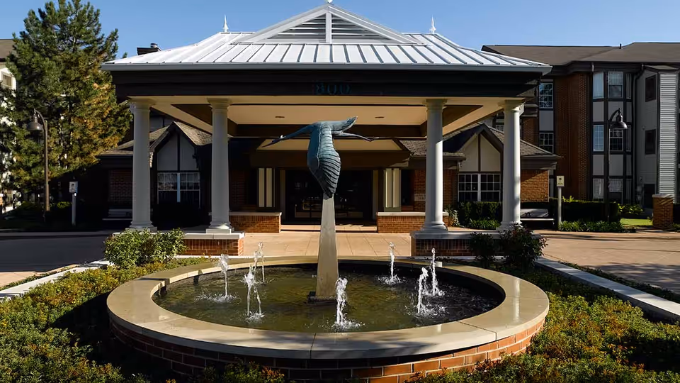 Front entrance of Atria Kinghaven facility featuring a covered driveway with white columns, a circular water fountain with a bird sculpture in the center, surrounded by greenery and shrubs, and the building facade in the background.