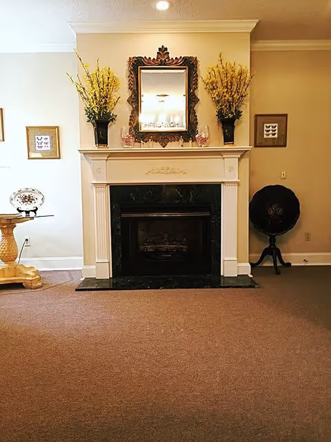 A cozy interior room featuring a white fireplace with a dark marble surround. Above the fireplace is an ornate framed mirror flanked by two vases filled with yellow flowers. On either side of the fireplace are framed butterfly pictures on the wall. To the left is a wooden table with a decorative plate, and to the right is a vintage-style black globe on a stand. The room has beige walls and carpeted flooring.