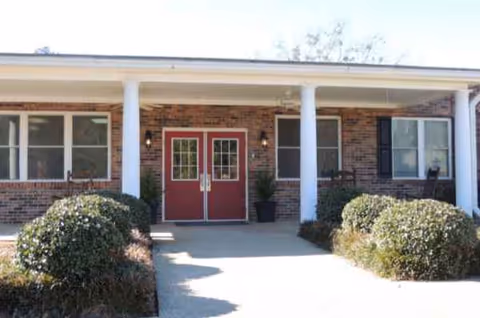 Brick nursing facility entrance with red double doors and manicured shrubs.