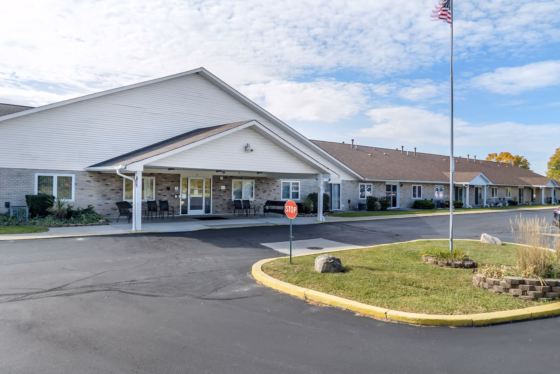 Exterior view of a single-story senior living facility building with a covered entrance, several chairs outside, a stop sign, an American flag on a flagpole, and a paved driveway with landscaped grassy areas.