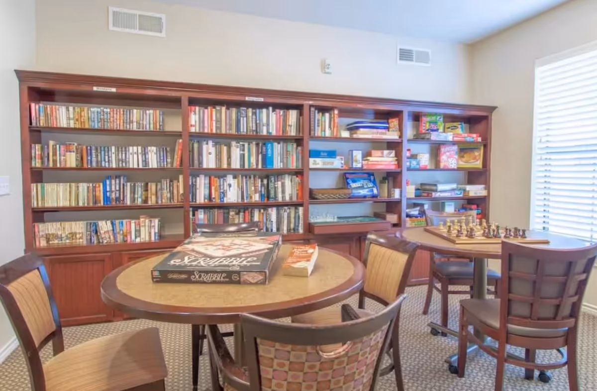 A cozy activity room with two round tables surrounded by chairs. One table has a Scrabble board game and a book on it, while the other table has a chessboard set up. In the background, there are large wooden bookshelves filled with books and various board games. A window with blinds allows natural light to brighten the room.