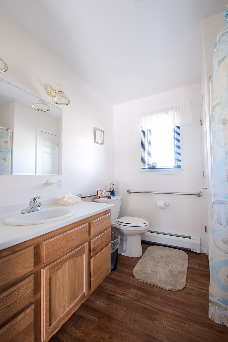 Bright residential bathroom with a wooden vanity and sink, toilet with grab bar, window, and shower curtain.