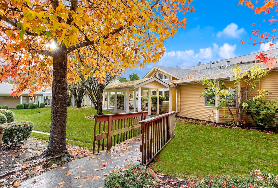 Outdoor view of a senior living facility with a wooden bridge over a small path, surrounded by green grass and trees with autumn-colored leaves. The building is light yellow with a covered walkway and a clear blue sky in the background.