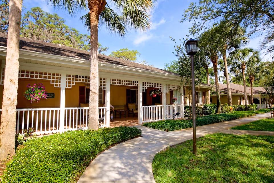 Front porch of a single-story cottage building with white railing, hanging flower baskets and a curved sidewalk amid palm trees.