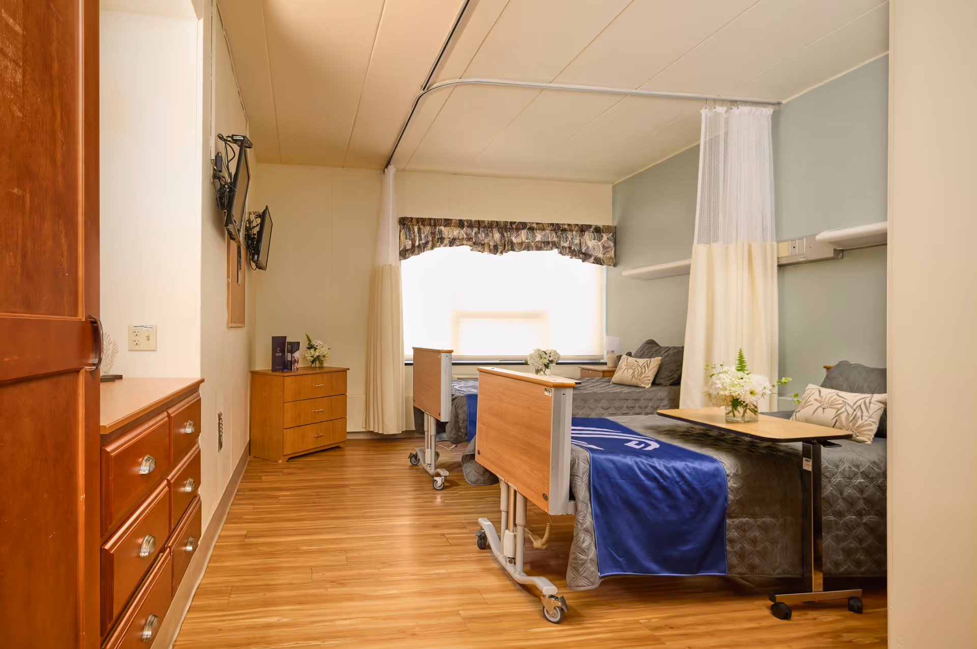 A bright and clean room with two hospital-style beds separated by a privacy curtain. Each bed has a gray quilted blanket and decorative pillows. There is a small wooden dresser and a mounted TV on the wall. The floor is wooden, and a large window with a valance lets in natural light. A small table with a flower arrangement is positioned next to one of the beds.
