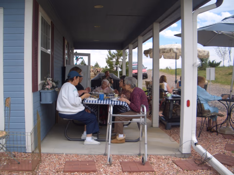 A group of elderly people sitting around a table with a blue and white checkered tablecloth on a covered porch outside a blue building, eating and socializing. There are additional people seated at tables with umbrellas in the background on a gravel area.