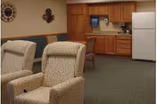 Common room with two upholstered recliners facing a small kitchenette with wooden cabinets and a white refrigerator.