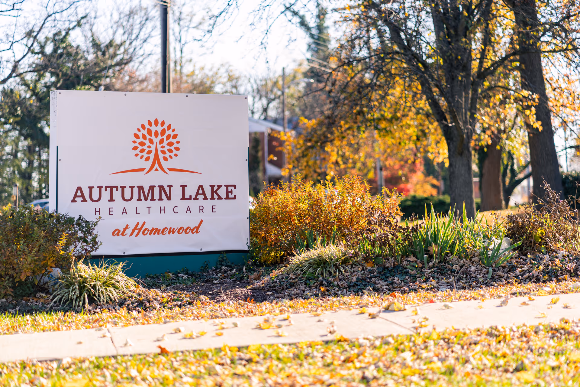 Outdoor view of a sign for Autumn Lake Healthcare at Homewood surrounded by bushes and trees with autumn foliage.