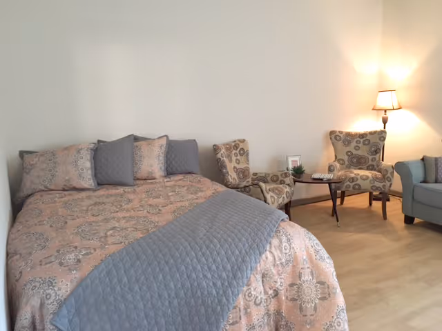 A cozy bedroom featuring a bed with a patterned pink and gray quilt and matching pillows. Next to the bed are two patterned armchairs with a small round table between them, holding a small plant and a framed photo. A floor lamp provides warm lighting, and a gray sofa is partially visible on the right side.