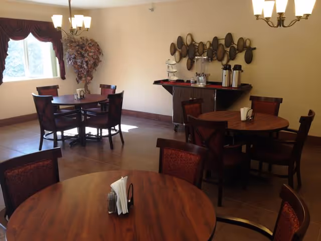 A dining room with round wooden tables and chairs with red upholstery. There is a window with a valance curtain, a decorative plant in the corner, a chandelier hanging from the ceiling, and a serving cart against the wall with coffee dispensers and cups. A decorative wall art piece is mounted above the cart.