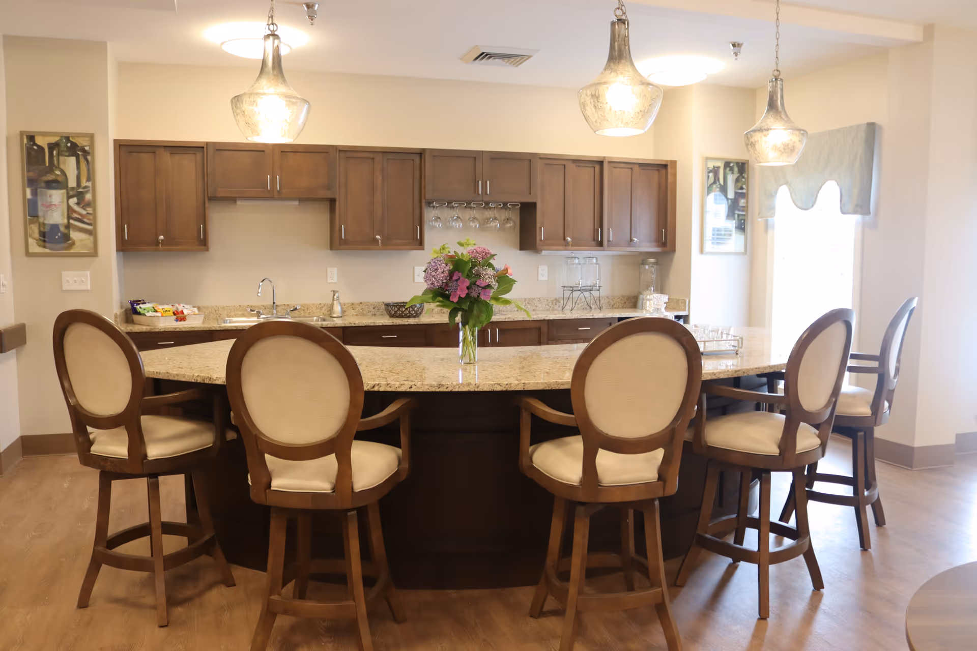 A bright kitchen area with a large granite island countertop surrounded by six wooden chairs with beige cushions. Above the island, there are three pendant lights. The back wall features wooden cabinets, a sink, and a rack with hanging wine glasses. A vase with purple and pink flowers is placed on the island. The room has light-colored walls and wooden flooring.