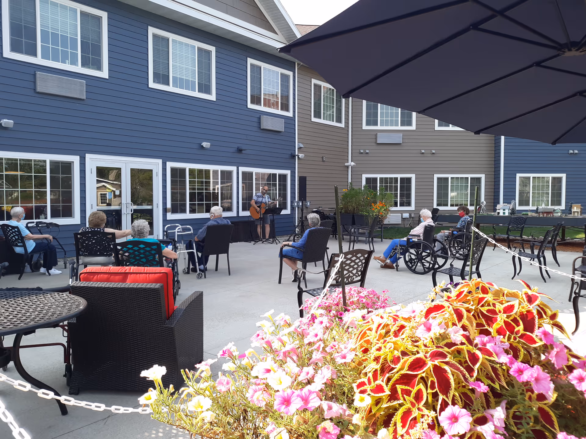 Outdoor patio area at a senior living facility with several elderly people seated in chairs and wheelchairs, socially distanced, watching a musician playing guitar and singing. The building exterior is visible with blue and beige siding and multiple windows. There are colorful flowers in the foreground and a large umbrella providing shade.