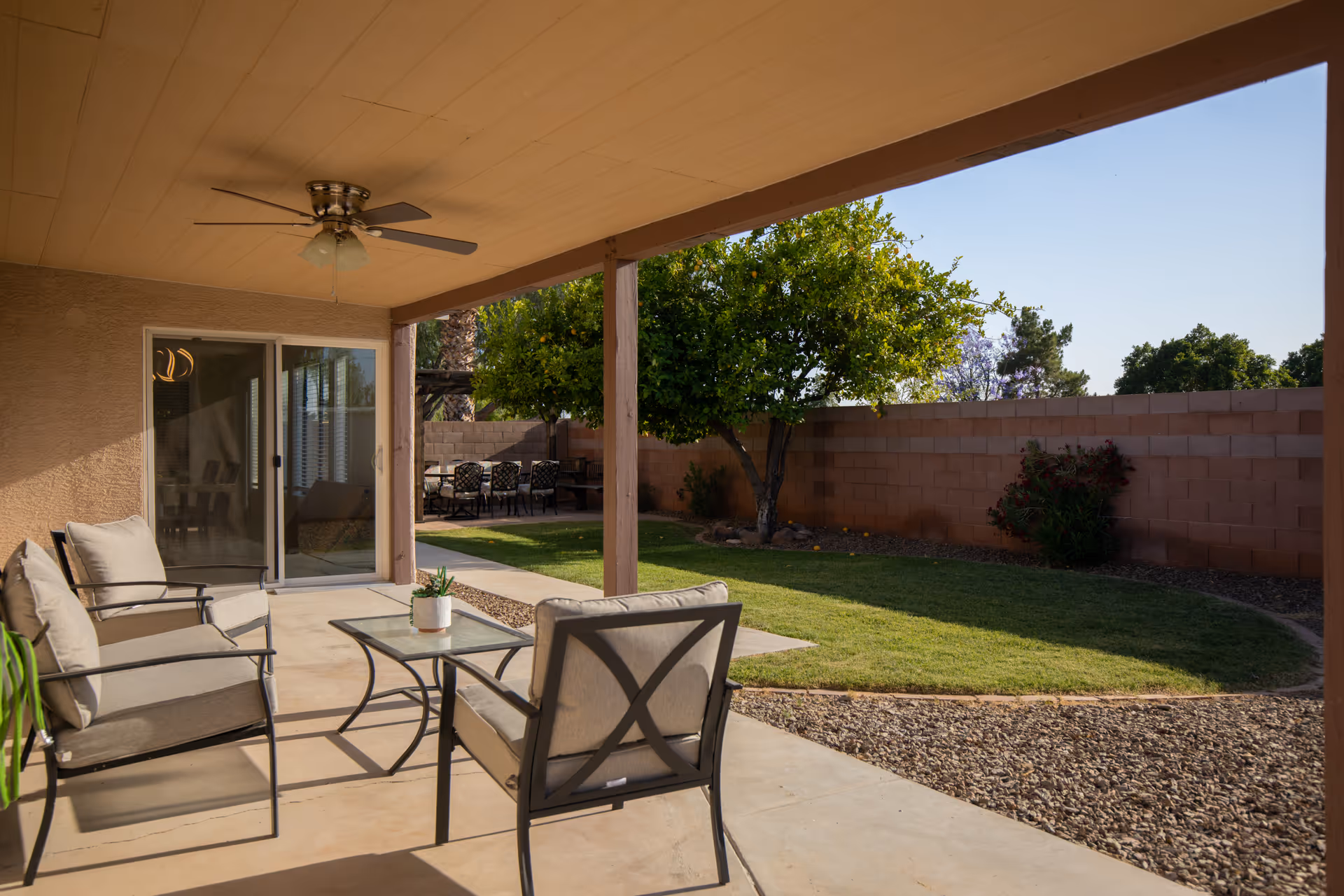 Covered patio with cushioned chairs and a glass-top table overlooking a grassy backyard with a tree and brick wall.