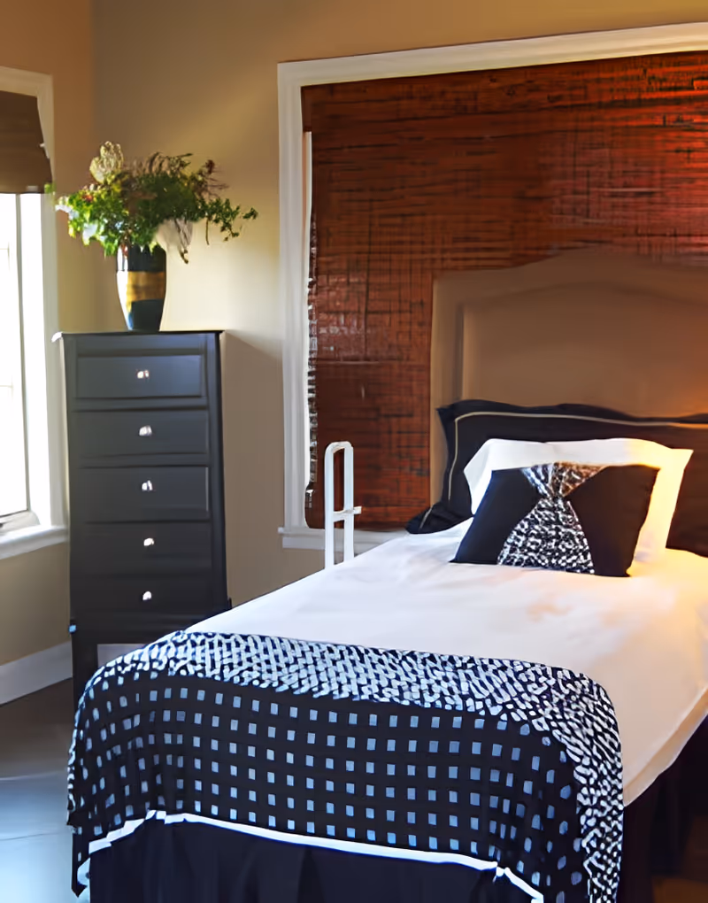 A neatly made single bed with a beige headboard, white sheets, and black and white patterned pillows and blanket. Next to the bed is a black chest of drawers with a vase of flowers on top, and a window with brown blinds partially visible.